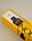 A sleek yellow box containing a bottle of Noén Nile Oil, a luxurious afterplay oil. The intimate oil is housed in a minimalist glass bottle with a wand applicator, making it easy to use. This indulgent product is formulated with a blend of natural ingredients, including beta-caryophyllene, calendula, moringa, buriti fruit, and rosemary, to soothe, rejuvenate, and revitalize the skin after sex.