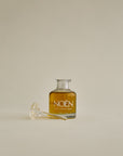 A glass bottle of Noén Nile Afterplay Oil, a luxurious afterplay oil designed to soothe intimate skin. The oil is housed in a glass bottle with a wand applicator, making it easy to use. The product features a blend of natural ingredients, including beta-caryophyllene, calendula, moringa, buriti fruit, and rosemary, to soothe, rejuvenate, and revitalize skin irritated after sex.