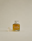 A glass bottle of Noén Nile Oil, a luxurious afterplay oil designed to soothe intimate skin. The oil is housed in a glass bottle with a wand applicator, making it easy to use. The product features a blend of natural ingredients, including beta-caryophyllene, calendula, moringa, buriti fruit, and rosemary, to soothe, rejuvenate, and revitalize skin irritated after sun, after sex, or after shower.