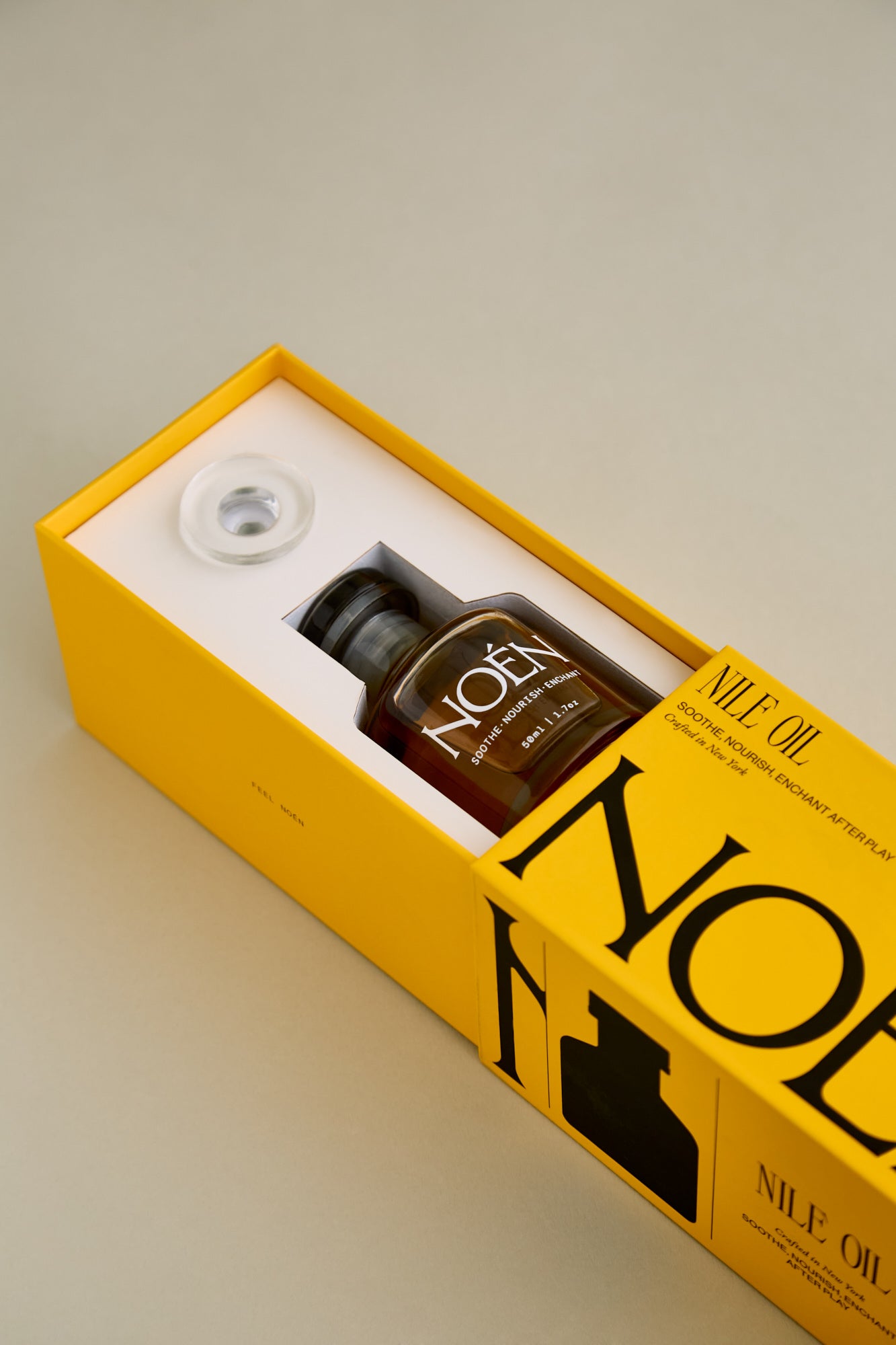 A sleek yellow box containing a bottle of Noén Nile Oil, a luxurious afterplay oil. The intimate oil is housed in a minimalist glass bottle with a wand applicator, making it easy to use. This indulgent product is formulated with a blend of natural ingredients, including beta-caryophyllene, calendula, moringa, buriti fruit, and rosemary, to soothe, rejuvenate, and revitalize the skin after sex.