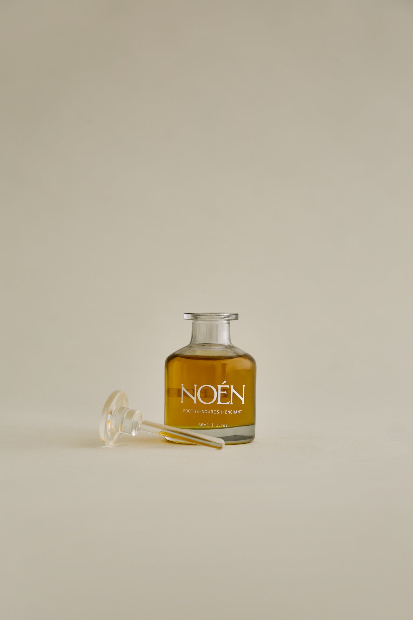 A glass bottle of Noén Nile Afterplay Oil, a luxurious afterplay oil designed to soothe intimate skin. The oil is housed in a glass bottle with a wand applicator, making it easy to use. The product features a blend of natural ingredients, including beta-caryophyllene, calendula, moringa, buriti fruit, and rosemary, to soothe, rejuvenate, and revitalize skin irritated after sex.